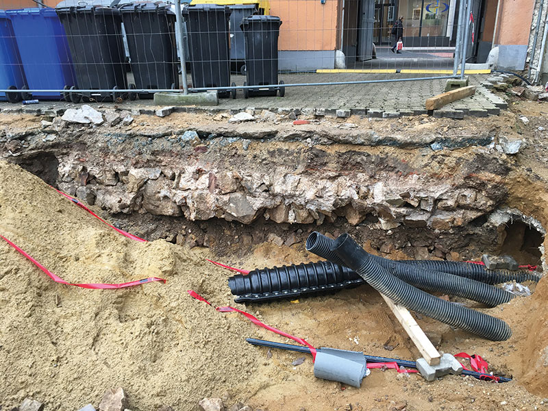 Nach dem Essen statten die fünf Museumsleute der Baustelle einen Besuch ab. Wann sieht man schon mal diese alte Mauer im Original? Die neuen Leitungen für das Kreishaus haben das Kastell durchschnitten, es liegt direkt unter unseren Füßen. Hinter dem Tor, auf der Düsseldorfer Straße, sieht man den Verlauf durch einen grauen Pflasterstreifen markiert.